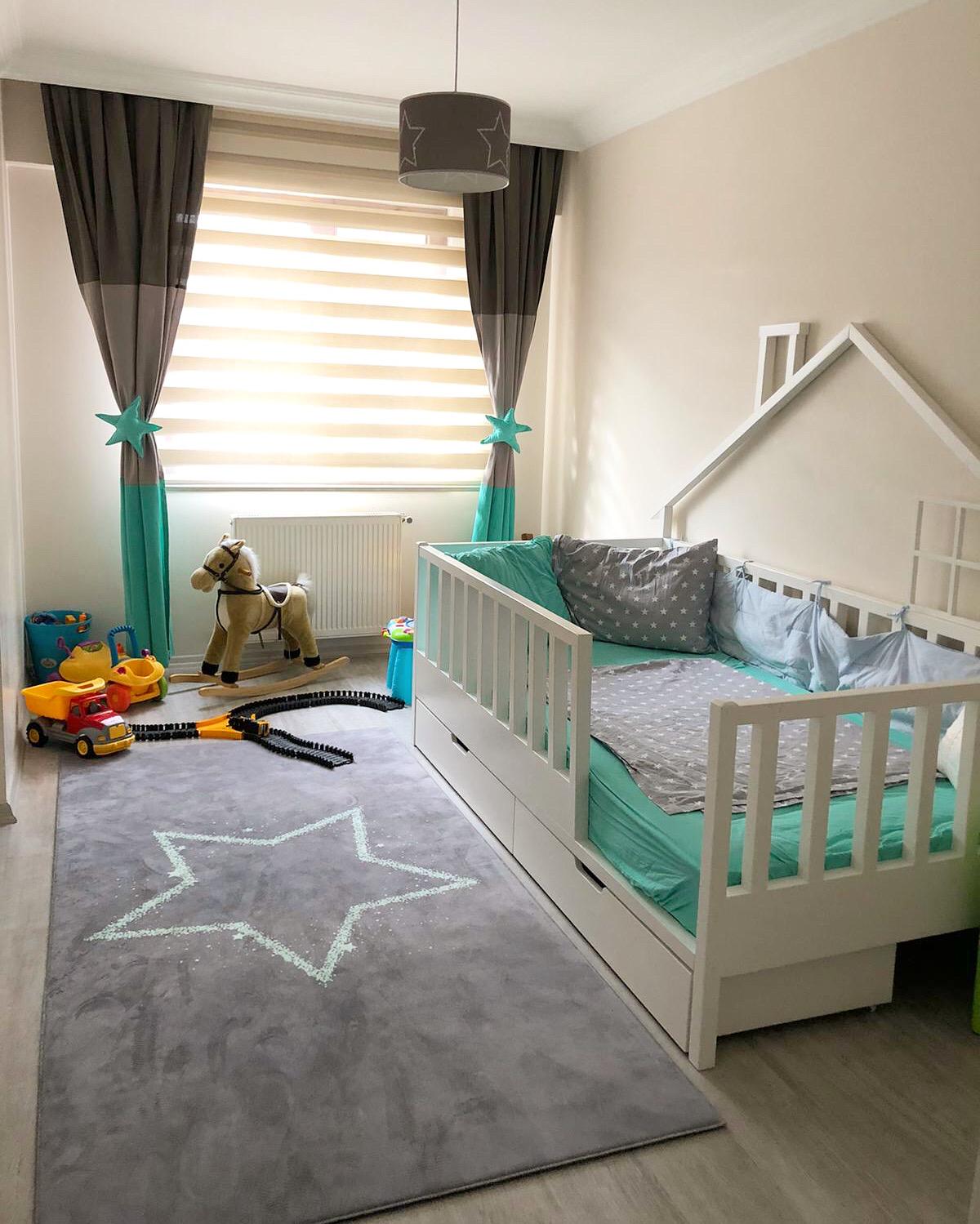 Halı Vagonu HV375 Mint Yıldızlı Gri çocuk odası halısı ile dekore edilmiş, soft renklerdeki bir çocuk odası. Odanın köşesinde beyaz bir beşikte mavi yastıklar, yan tarafında oyuncaklar ve odanın ortasında mavi yıldız desenli halı yer alıyor.
