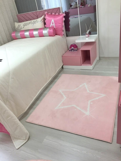 Pembe beyaz yıldız deseniyle süslenmiş çocuk halısı, modern bir odada yerleştirilmiş. Halının etrafında pembe ve beyaz dekoratif eşyalar, rahat bir yatak ve şık bir oda düzeni gözükmekte.