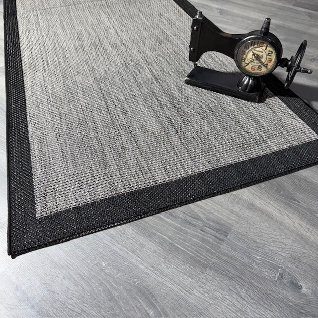 Gri ve siyah desenli jüt sisal halının kenarı, modelin altına konmuş vintage saat.