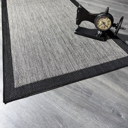 Gri ve siyah desenli jüt sisal halının kenarı, modelin altına konmuş vintage saat.