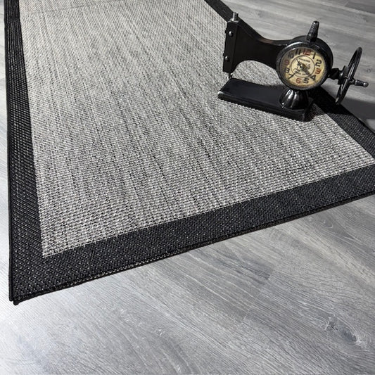 Gri ve siyah desenli jüt sisal halının kenarı, modelin altına konmuş vintage saat.